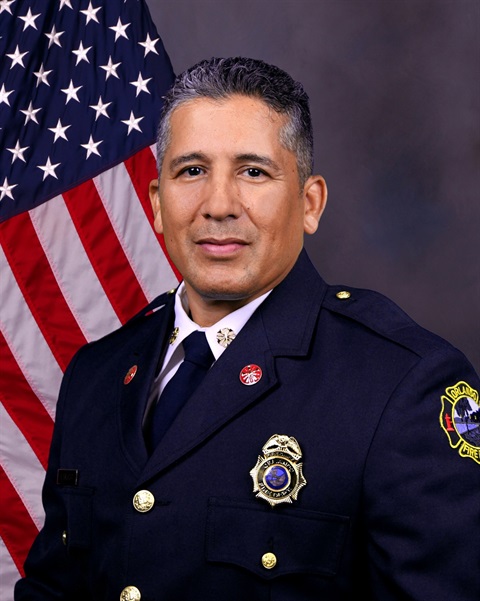 OFD Fire Chief Charles Salazar sits in front of a flag and wears his uniform jacket and hat
